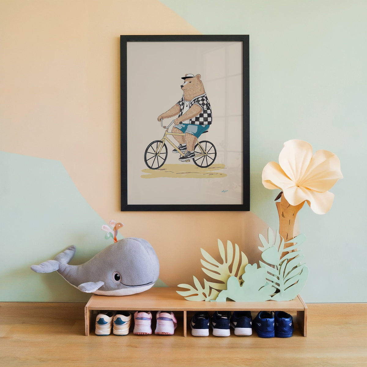 Brown bear riding yellow bicycle nursery print in a black frame on a mint and peach wall above a whale plush