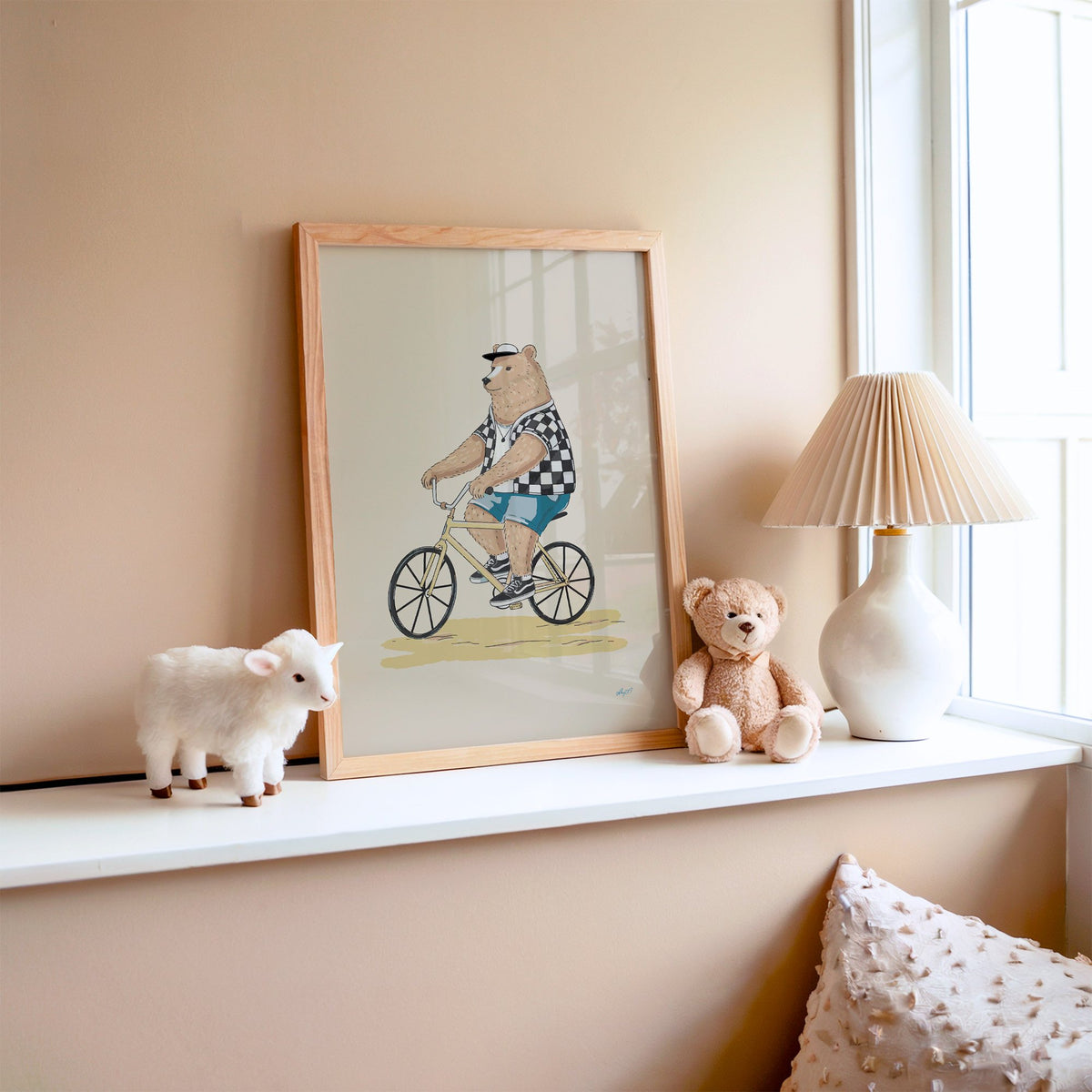 Brown bear riding yellow bicycle nursery print in an oak frame on a window ledge with a toy lamb and teddy
