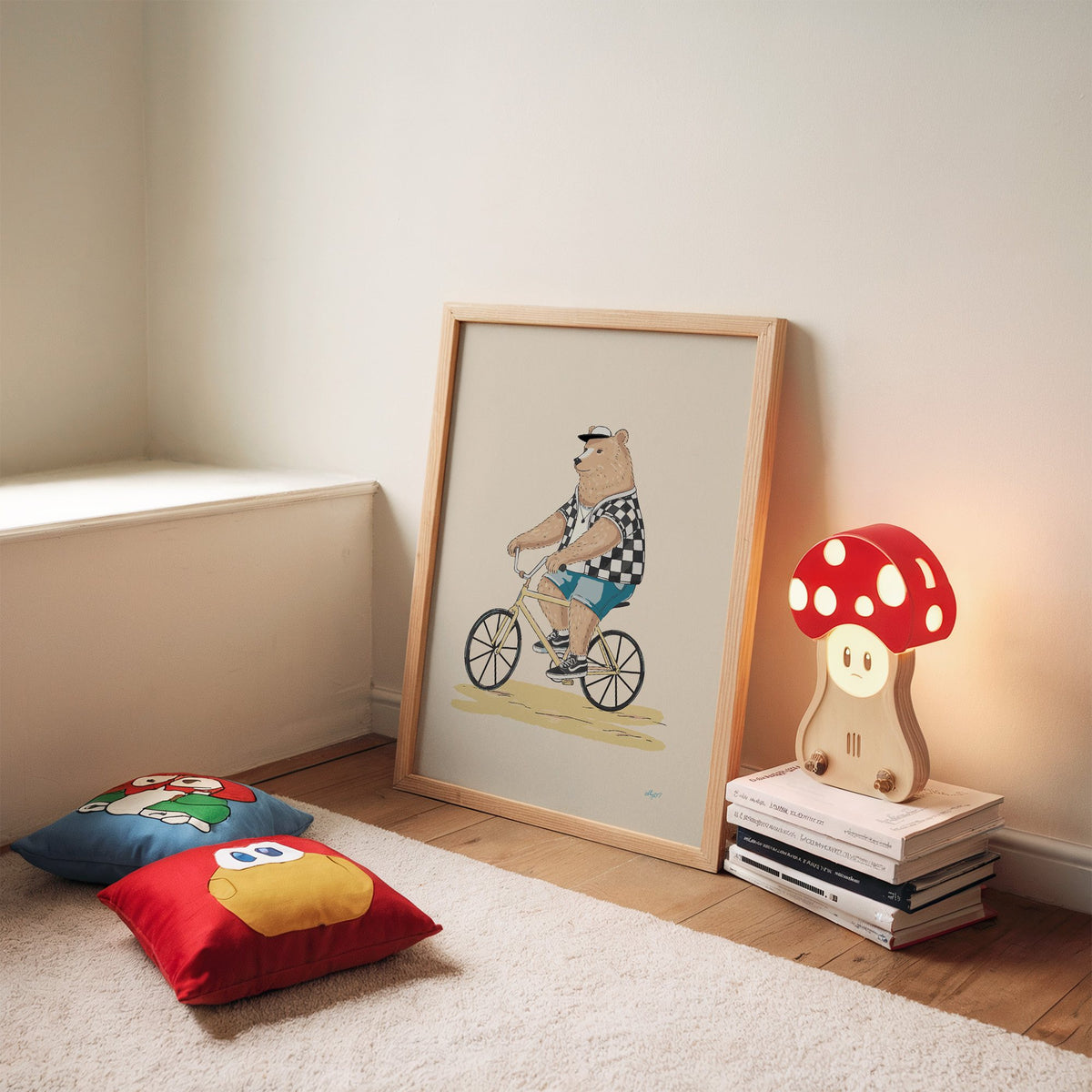 Brown bear riding yellow bicycle nursery print in an oak frame leaning on a rug beside a mushroom lamp and floor cushions