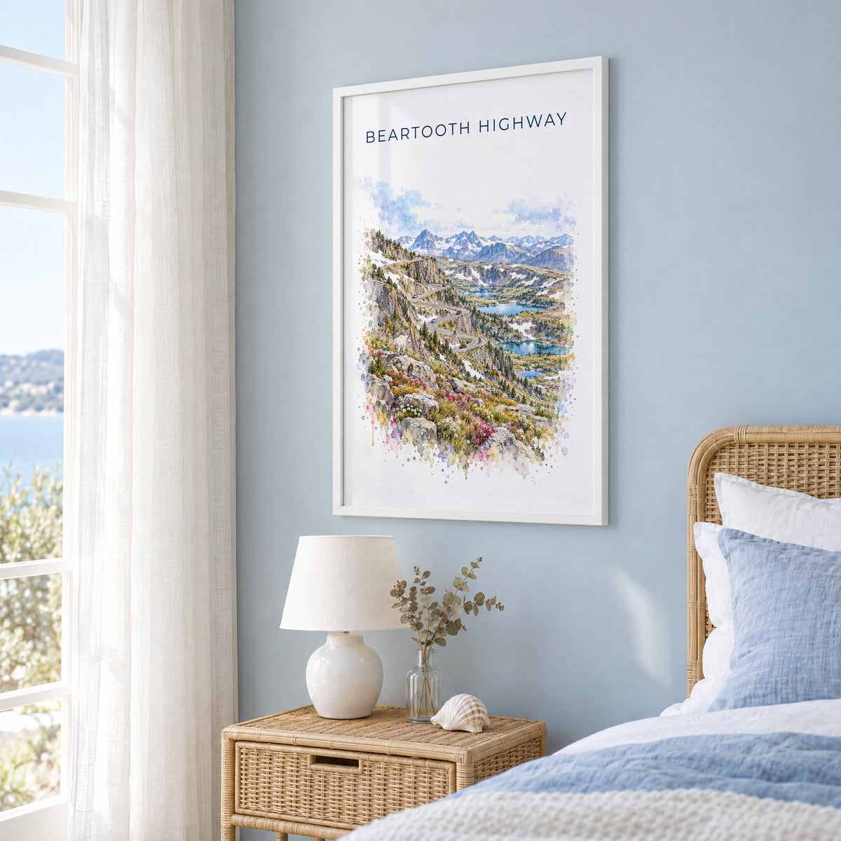 Beartooth Highway watercolor print in a white frame on a pale blue wall beside a rattan bed in a coastal bedroom