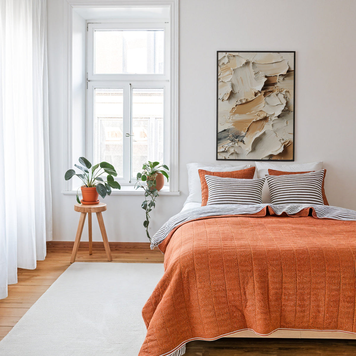 Beige and brown impasto texture abstract print in a thin black frame above a bed with a rust orange quilt