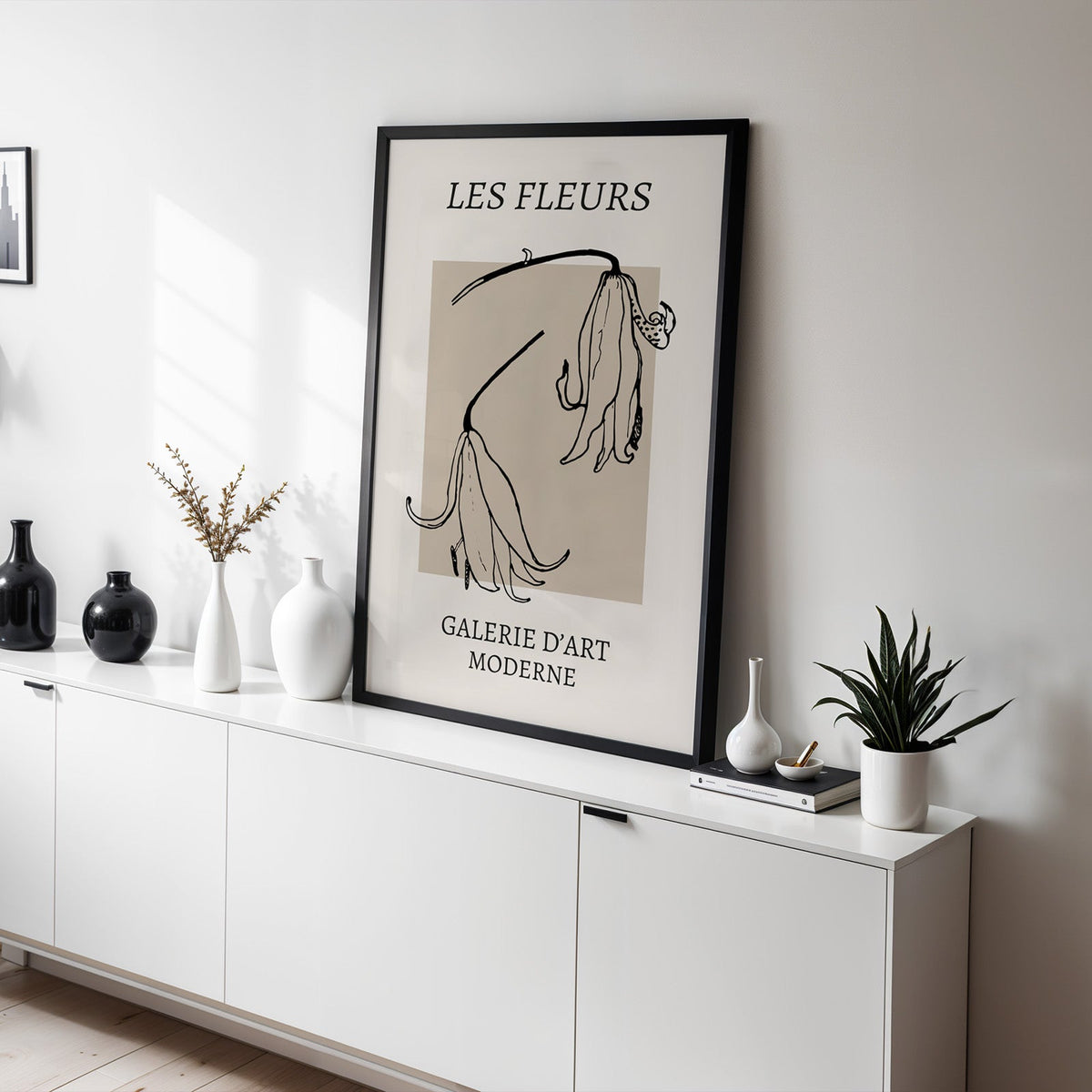 Drooping lily sketch on beige block botanical print in a black frame leaning on a white sideboard with black and white vases