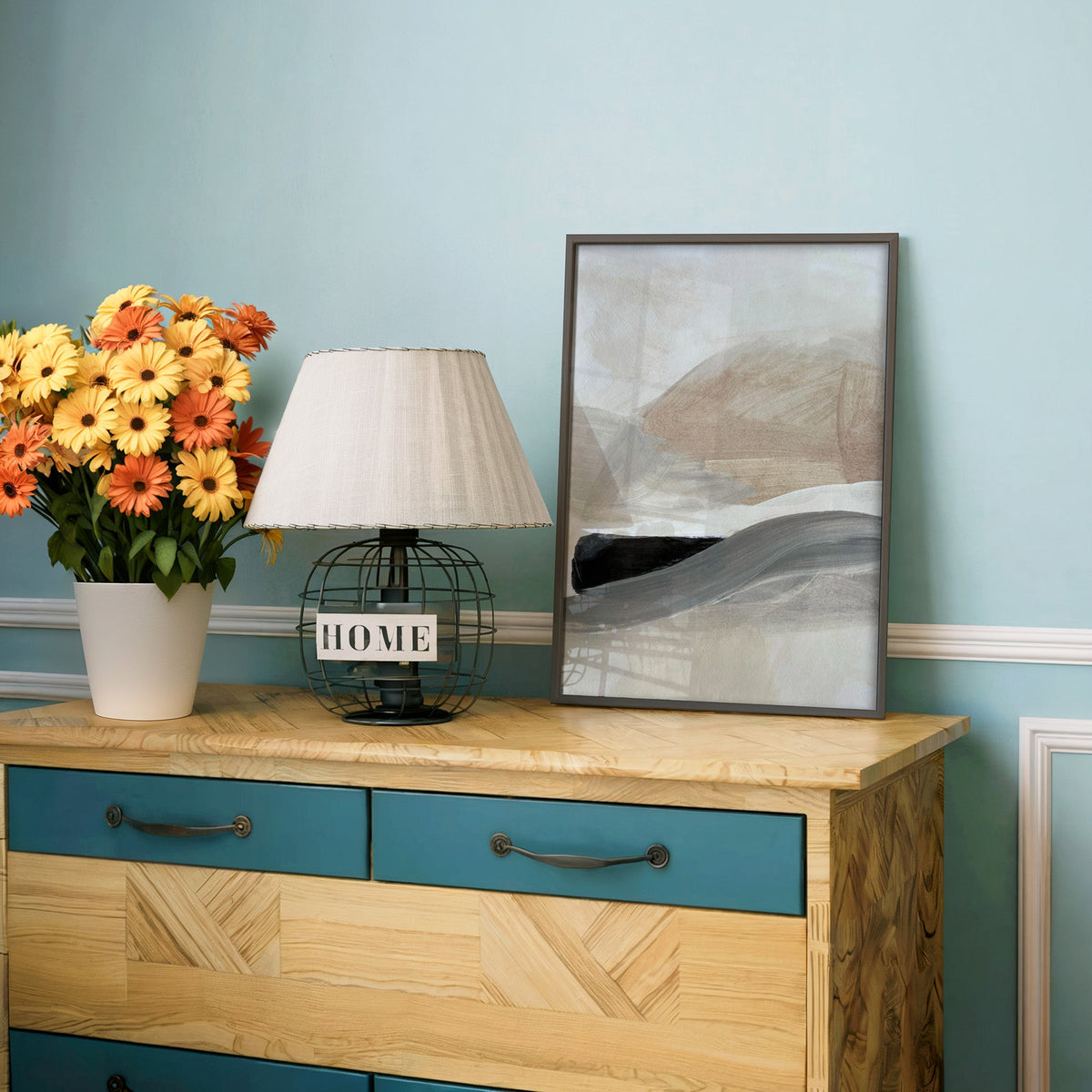 Beige hills and gray black wave abstract print in a charcoal frame on a teal dresser beside orange and yellow daisies