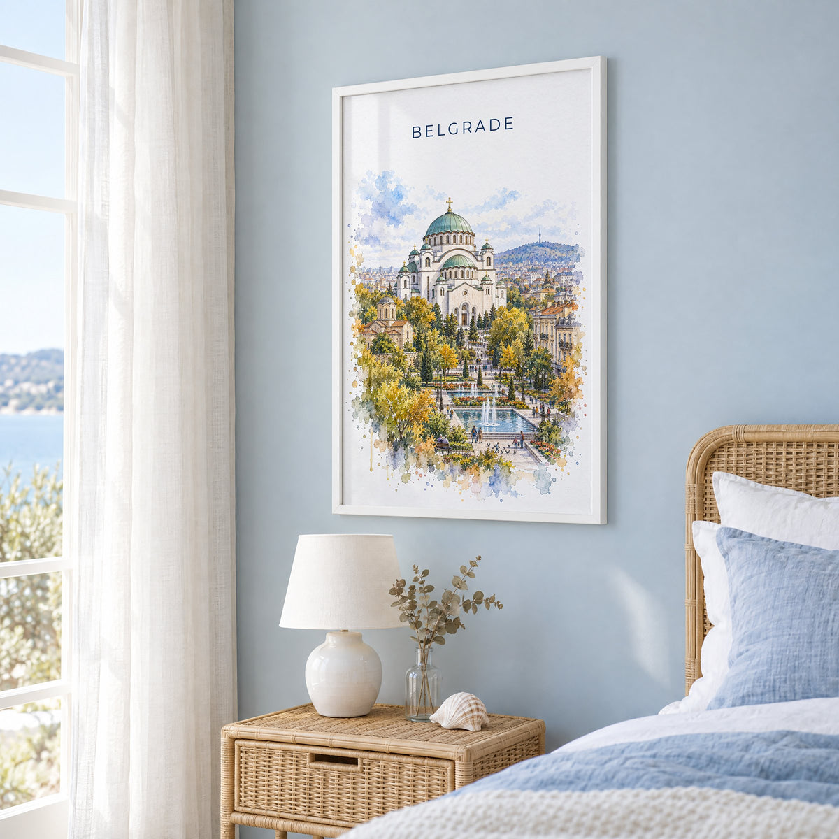Belgrade watercolor print in a white frame on a pale blue wall beside a rattan bed in a coastal bedroom