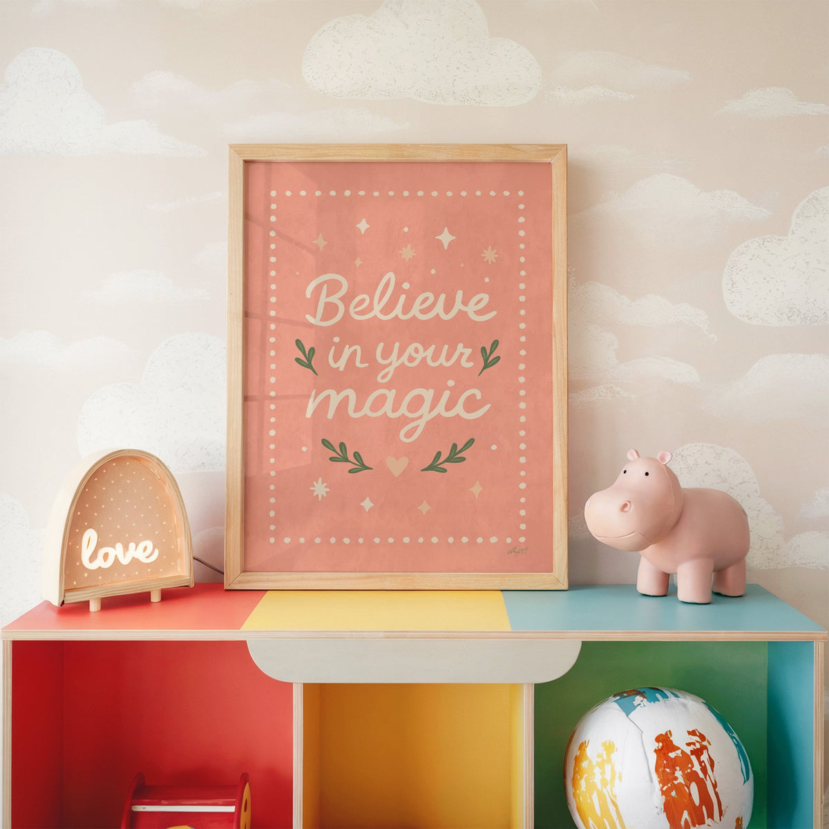 Believe in your magic coral quote nursery print in an oak frame on a rainbow toy shelf against cloud wallpaper