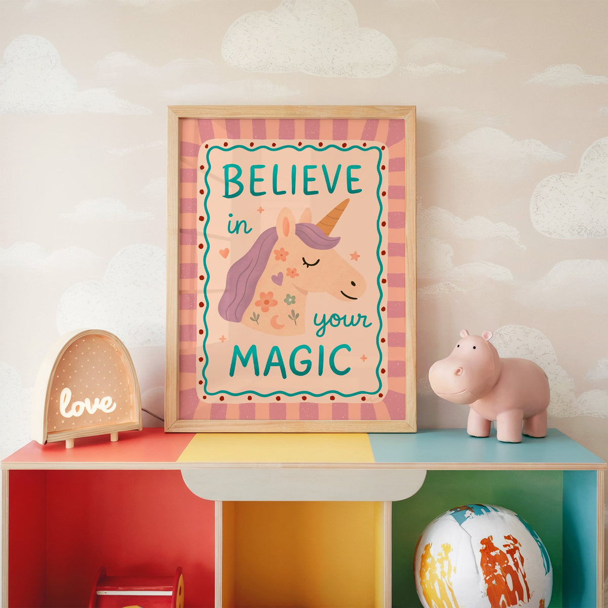 Believe in your magic unicorn nursery print in an oak frame on a rainbow toy shelf against cloud wallpaper