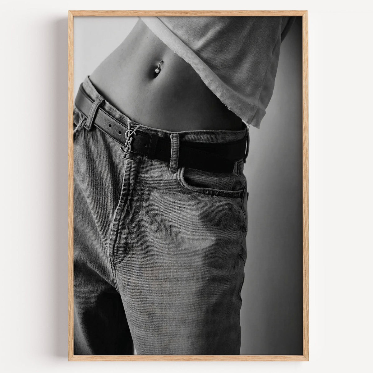 Belly piercing above faded jeans and belt buckle print in a light oak frame on a plain white wall