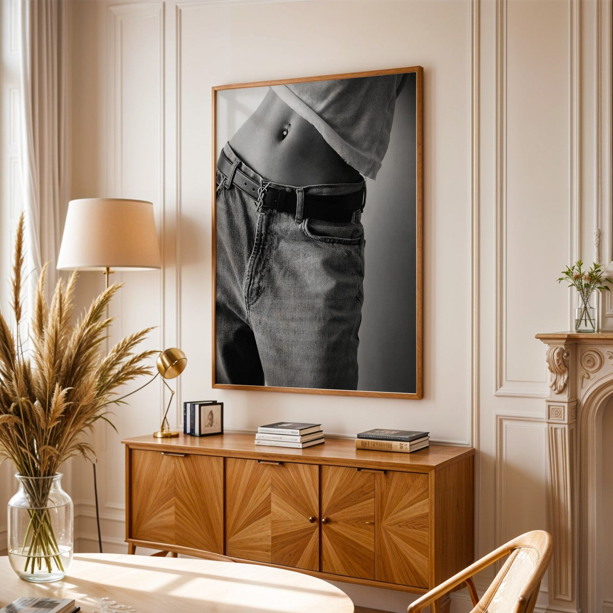 Belly piercing above faded jeans and belt buckle print in an oak frame above a sunburst wood sideboard with pampas grass