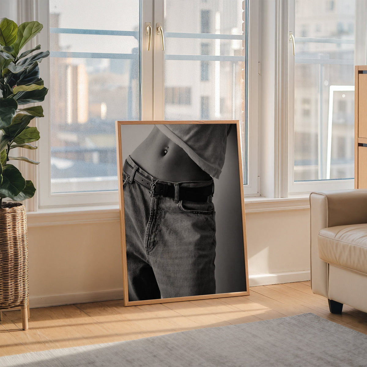 Belly piercing above faded jeans and belt buckle print in an oak frame leaning on the floor beside a fiddle leaf fig