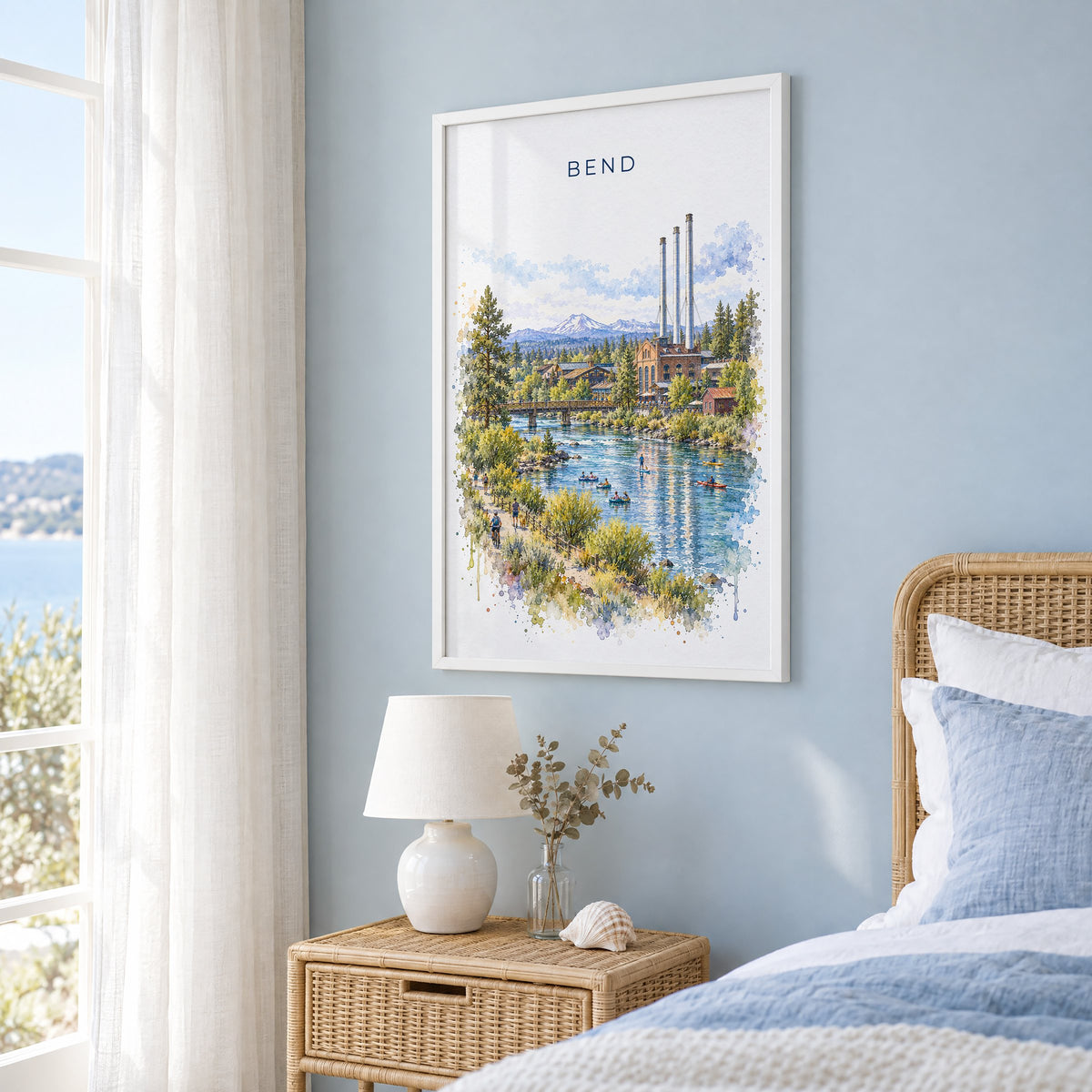 Bend, Oregon watercolor print in a white frame on a pale blue wall beside a rattan bed in a coastal bedroom