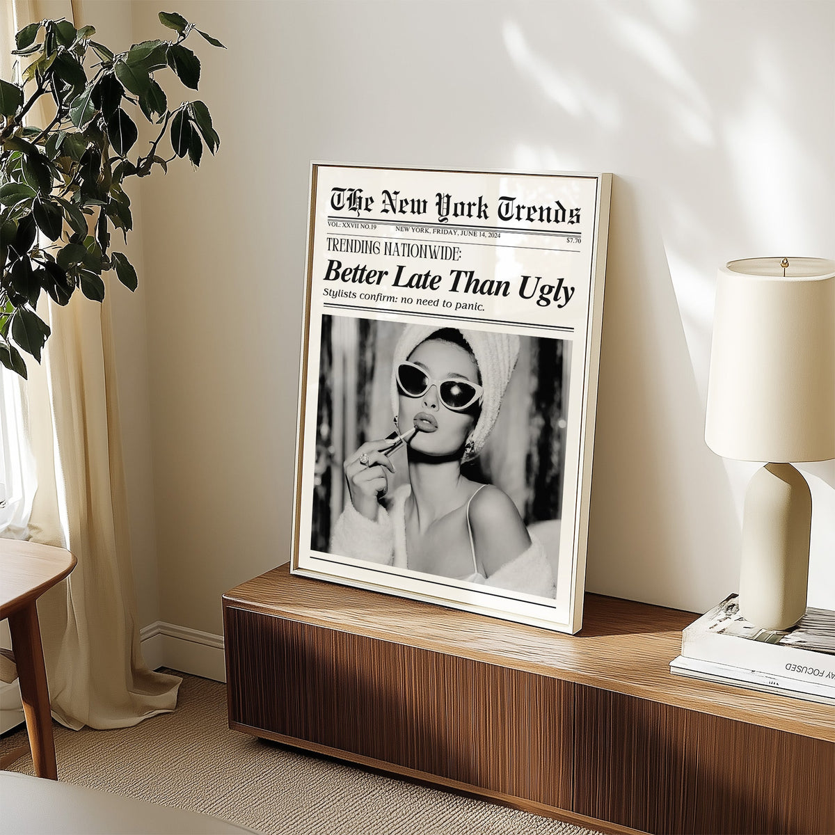 Woman applying lipstick mock newspaper print in a white frame on a walnut sideboard beside a ceramic lamp