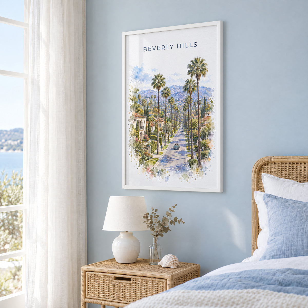 Beverly Hills watercolor print in a white frame on a pale blue wall beside a rattan bed in a coastal bedroom
