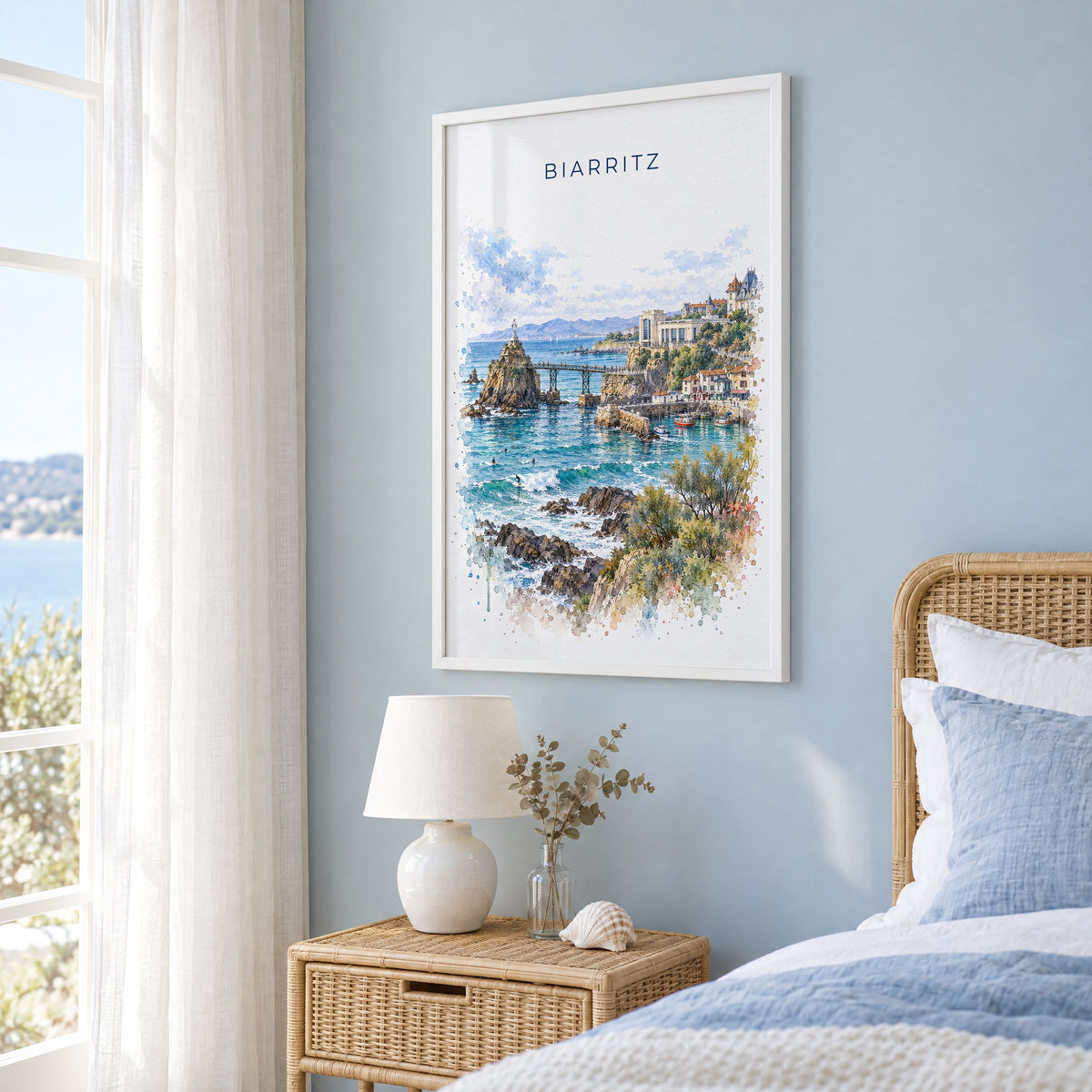 Biarritz watercolor print in a white frame on a pale blue wall beside a rattan bed in a coastal bedroom