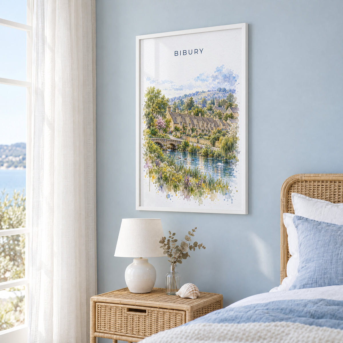 Bibury watercolor print in a white frame on a pale blue wall beside a rattan bed in a coastal bedroom
