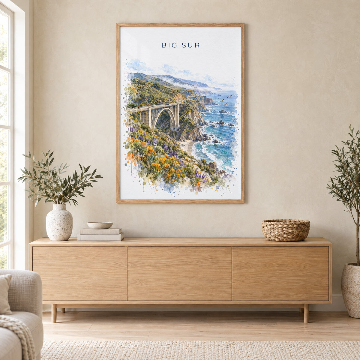 Big Sur watercolor print in an oak frame above a light wood sideboard in a Scandinavian living room