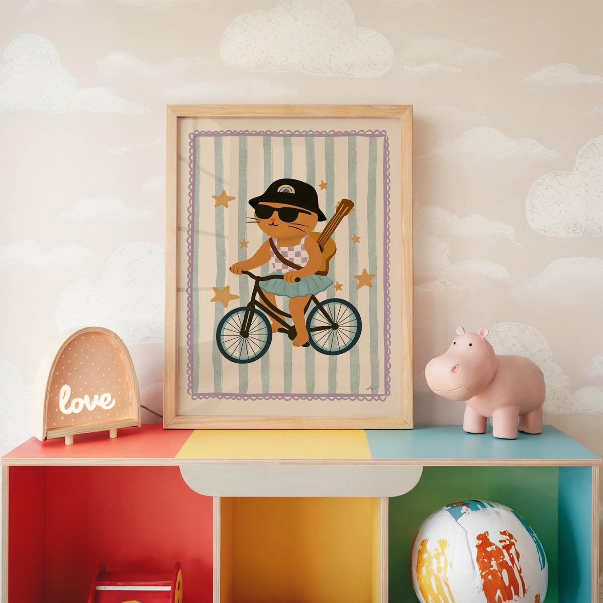 Orange cat on bicycle with ukulele nursery print in an oak frame on a rainbow toy shelf against cloud wallpaper