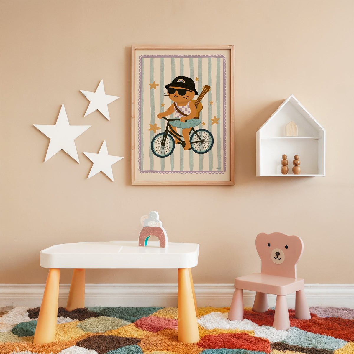 Orange cat on bicycle with ukulele nursery print in an oak frame on a peach wall above a kids table with wall stars