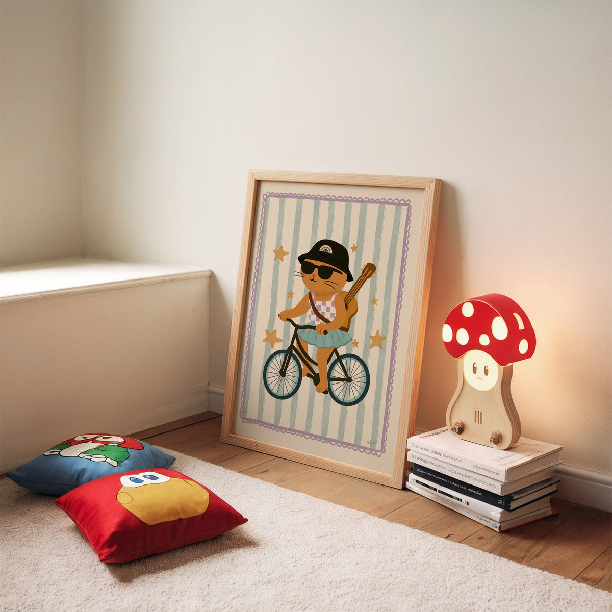 Orange cat on bicycle with ukulele nursery print in an oak frame leaning on a rug beside a mushroom lamp and floor cushions