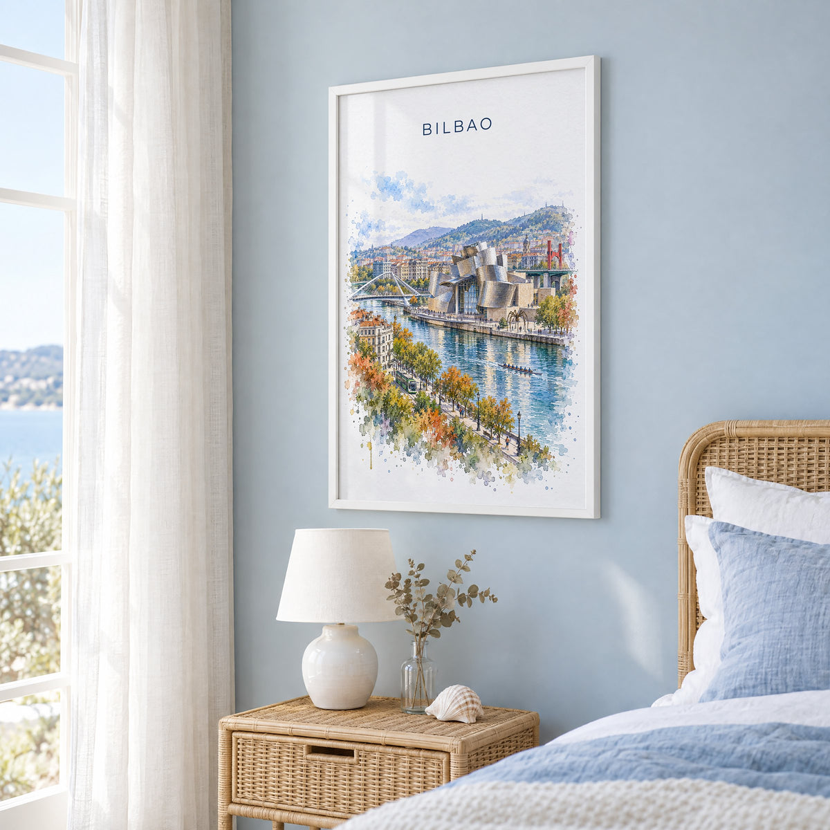 Bilbao watercolor print in a white frame on a pale blue wall beside a rattan bed in a coastal bedroom