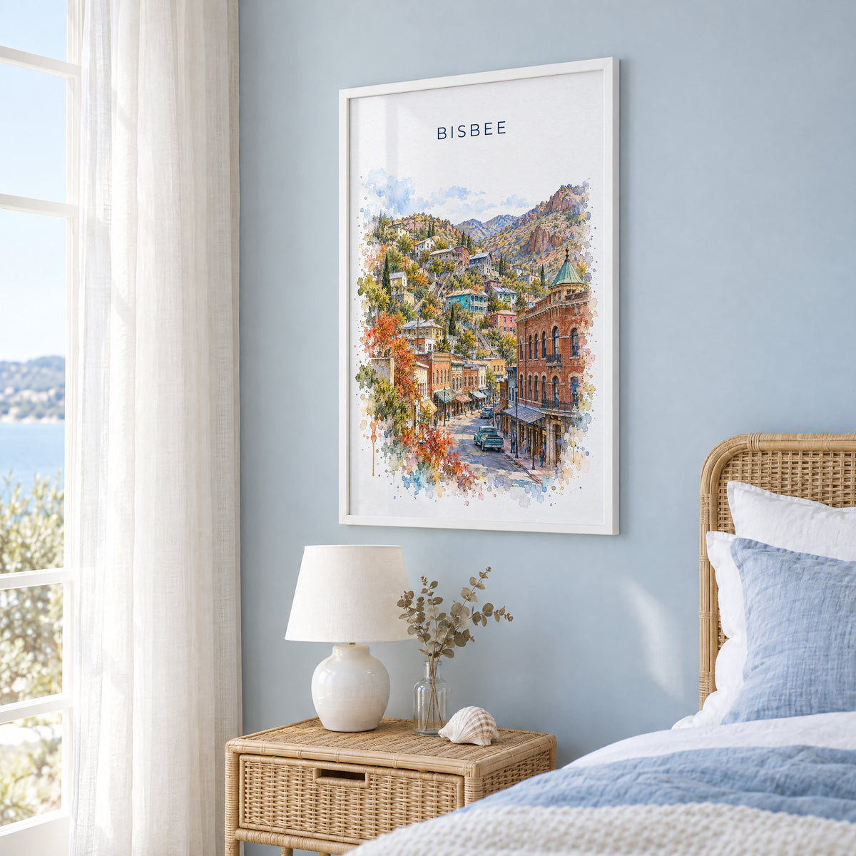 Bisbee watercolor print in a white frame on a pale blue wall beside a rattan bed in a coastal bedroom