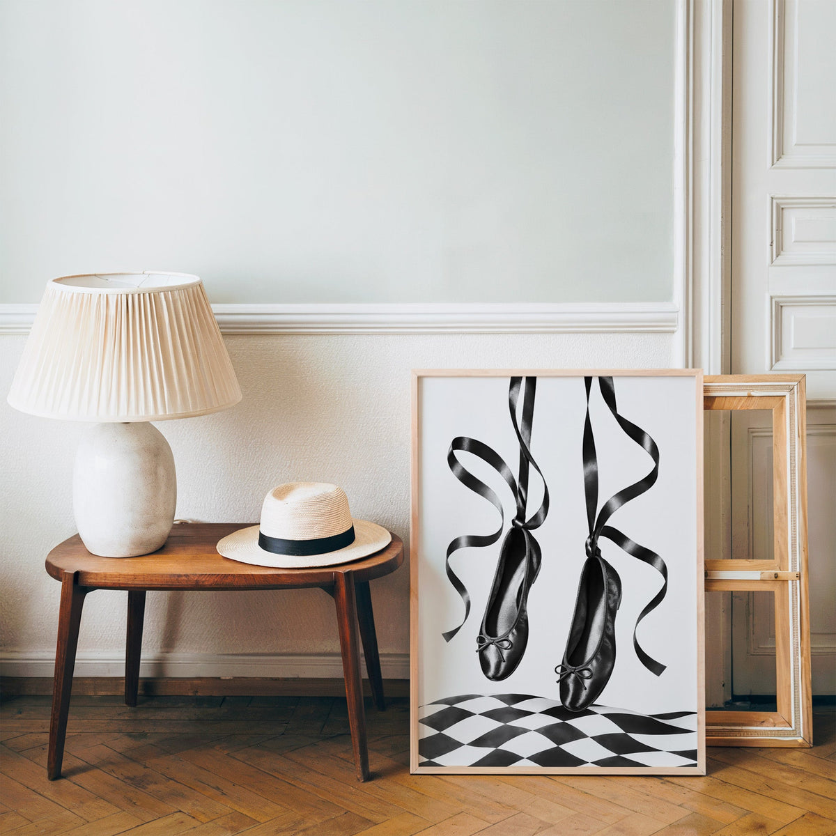 Black ballet flats on ribbons over checkerboard print in a pine frame on a wood floor beside a lamp and straw hat