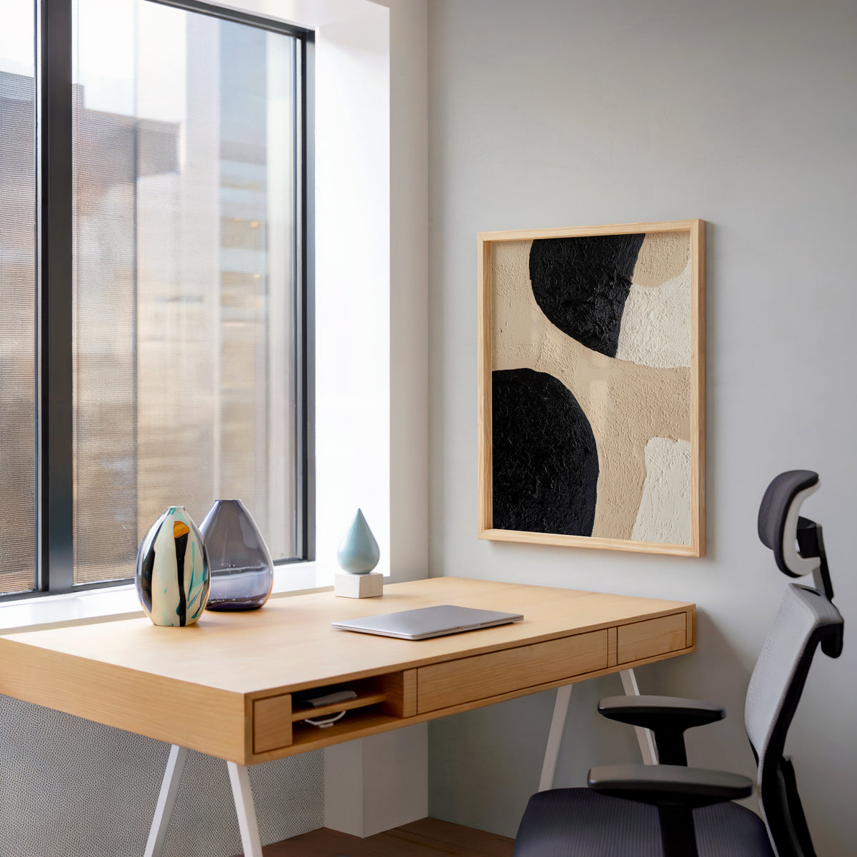Black and beige textured shapes abstract print in a pale wood frame above a home office desk by a tall window