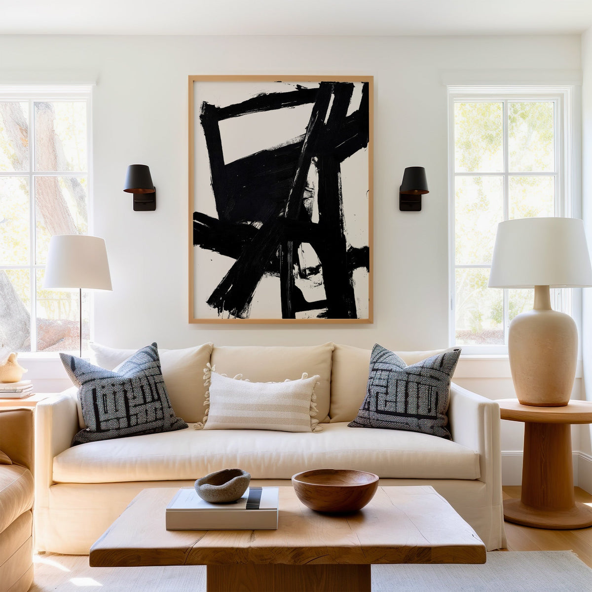Black bold crossbeam brushstrokes on white print in a light oak frame above a cream sofa between two black wall sconces