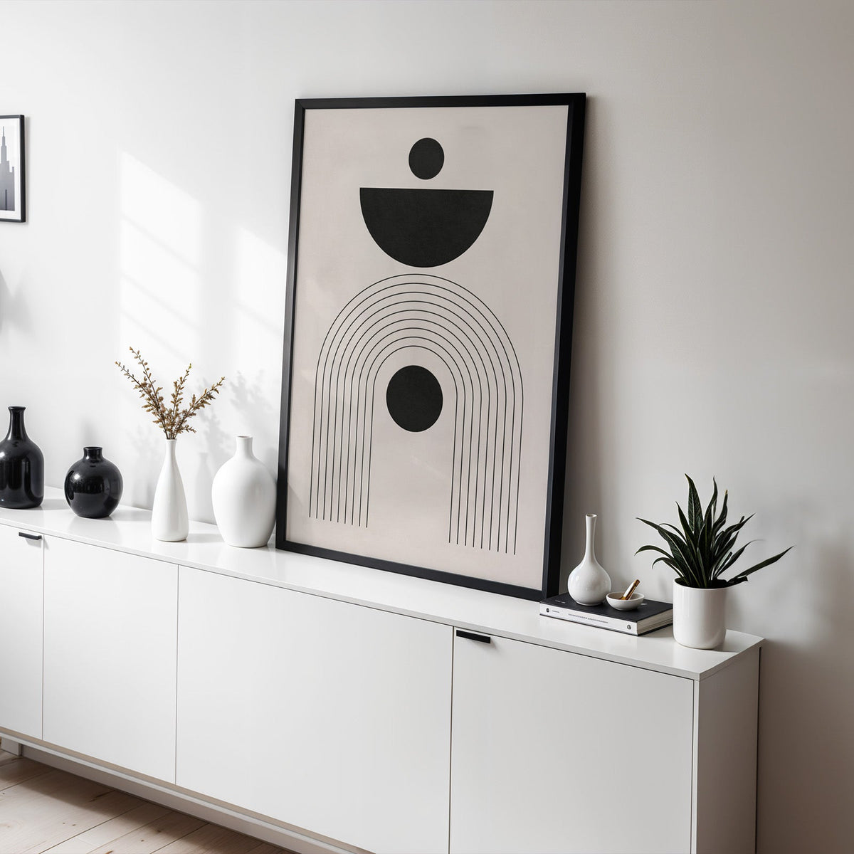 Black bowl and circle totem mid century print in a black frame leaning on a white sideboard with black and white vases