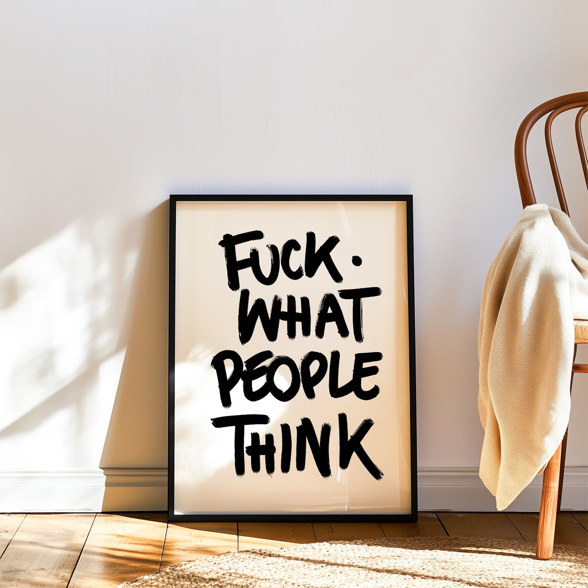Black brush lettering attitude quote print in a black frame leaning on a wood floor beside a bentwood chair