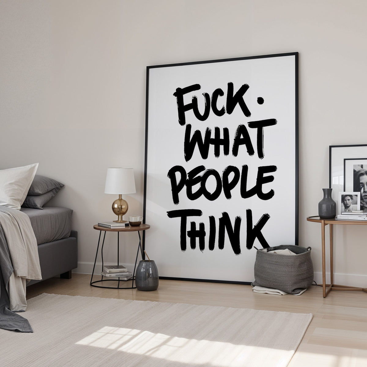 Black brush lettering attitude quote print in a black frame leaning in a bedroom beside a gold lamp