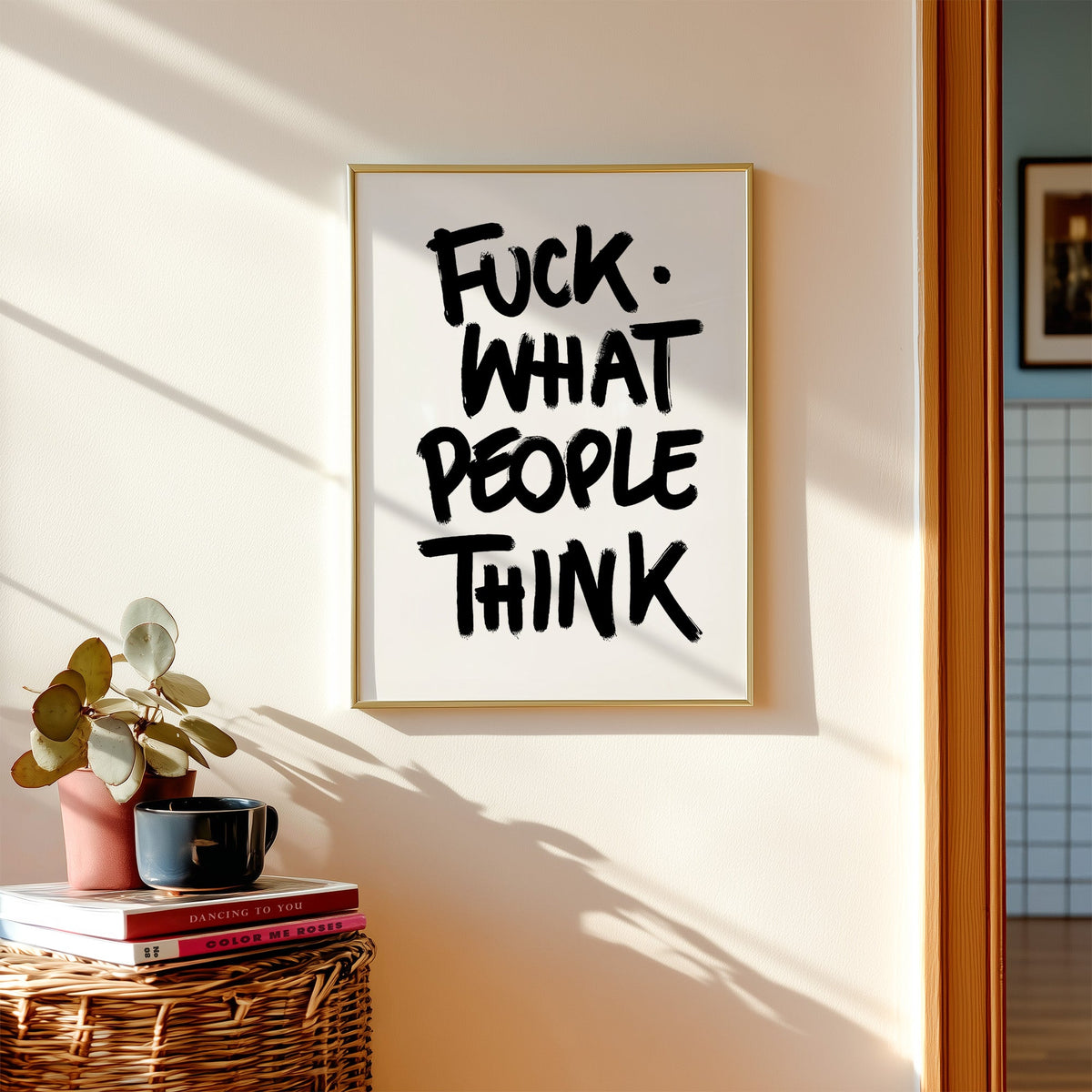 Black brush lettering attitude quote print shown in a gold frame above a wicker basket and stacked books