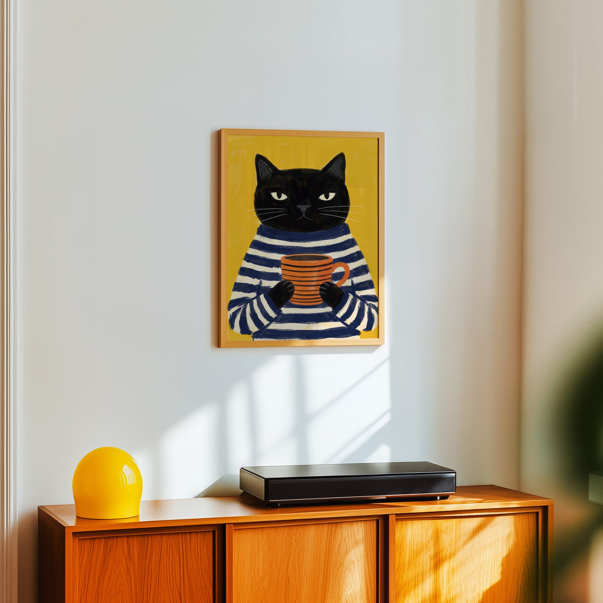 Yellow cat coffee print in a light oak frame above a teak sideboard with a yellow lamp