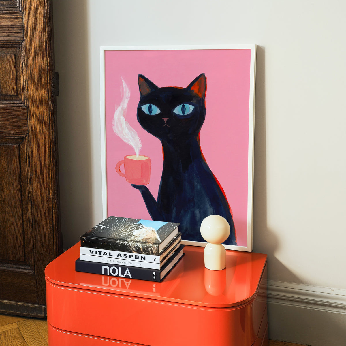 Cat holding coffee print in a white frame on a glossy orange cabinet with books