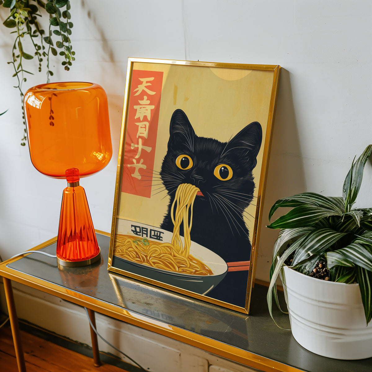 Black cat slurping ramen print shown in a gold frame on a console beside an orange glass lamp