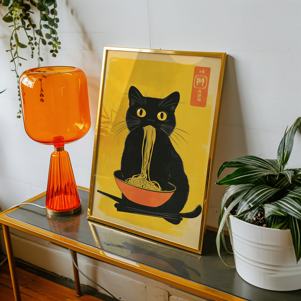 Black cat noodles orange bowl print shown in a gold frame on a console beside an orange glass lamp