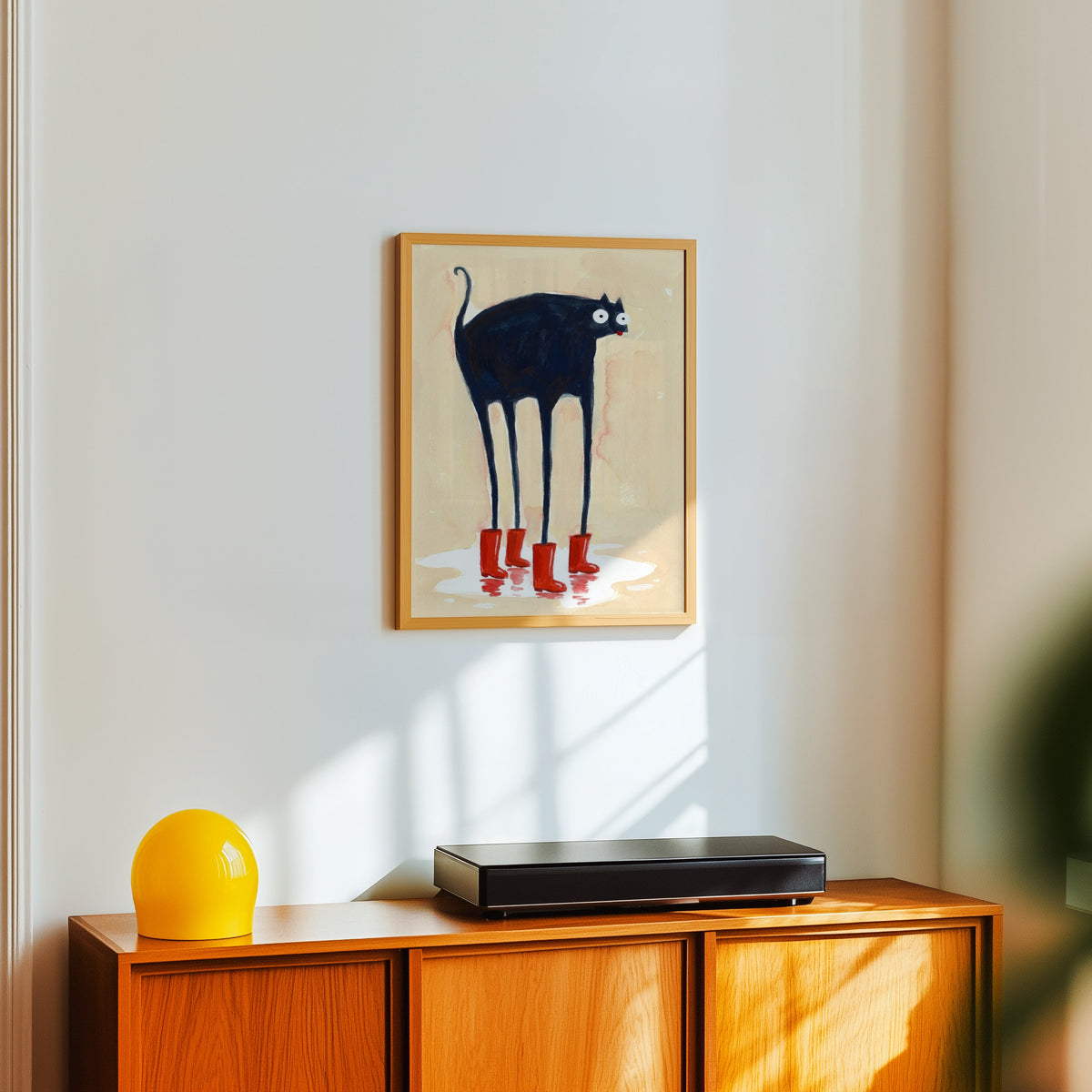 Red boots cat print in a light oak frame above a teak sideboard with a yellow lamp