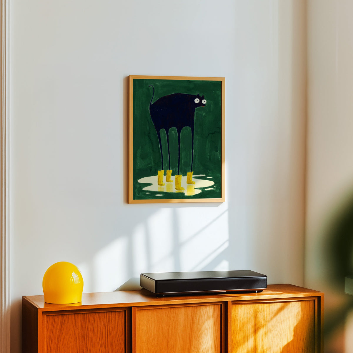 Googly-eyed black cat print in a light oak frame above a teak sideboard with a yellow lamp
