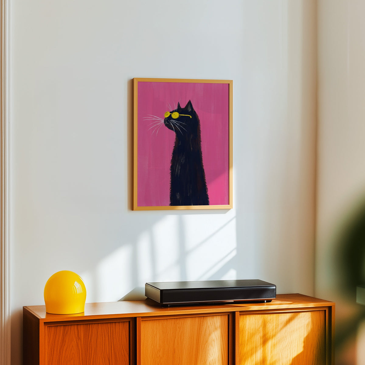 Fuchsia pink cat print in a light oak frame above a teak sideboard with a yellow lamp