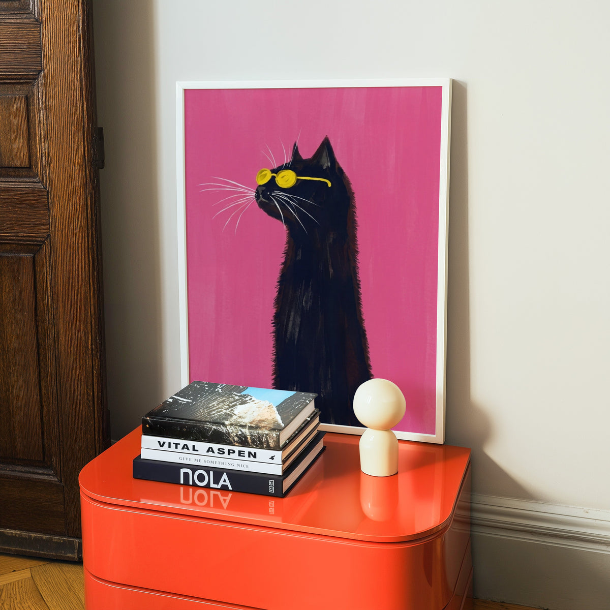 Cool cat on hot pink print in a white frame on a glossy orange cabinet with books