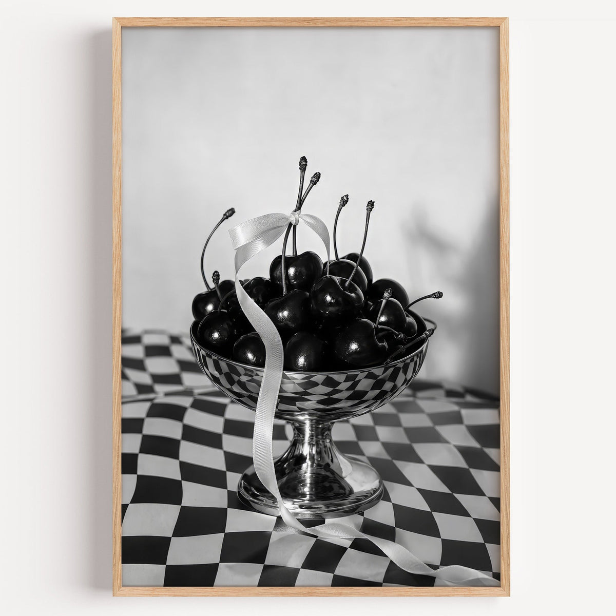 Black cherries in footed silver bowl print in a light oak frame on a plain white wall