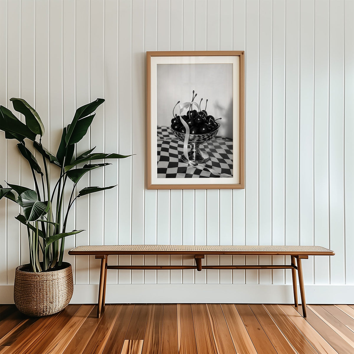 Black cherries in footed silver bowl print in a wood frame with a white mat above a cane bench