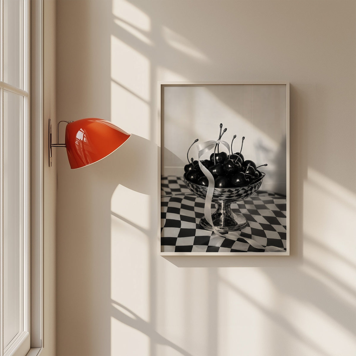 Black cherries in footed silver bowl print in a white frame on a sunlit wall beside a red sconce