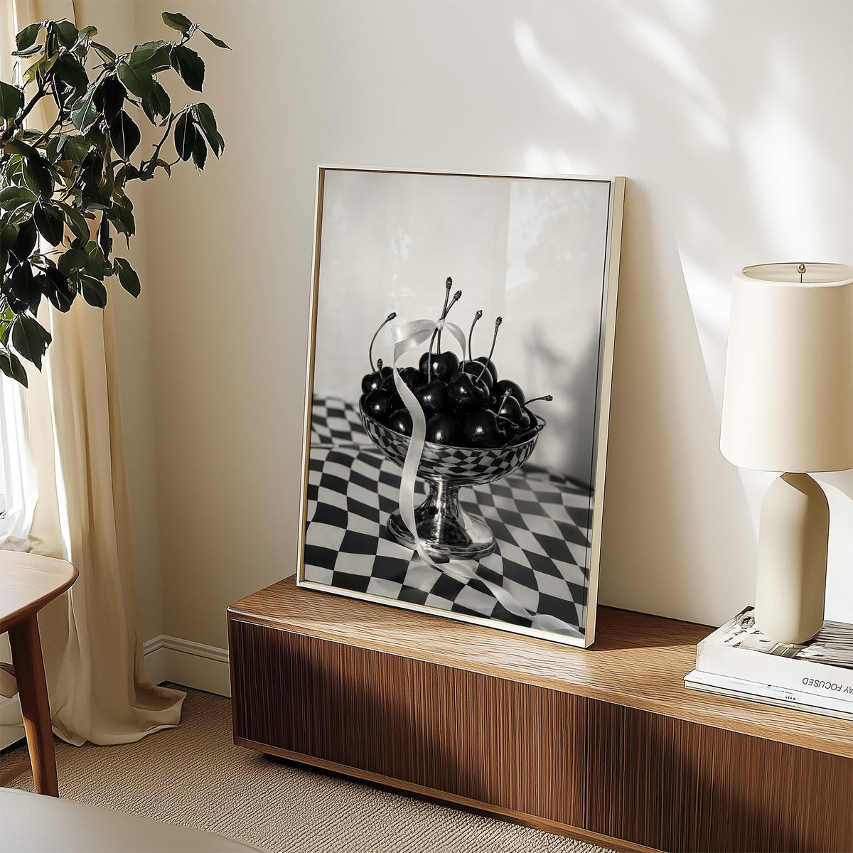 Black cherries in footed silver bowl print in a white frame on a walnut sideboard beside a ceramic lamp