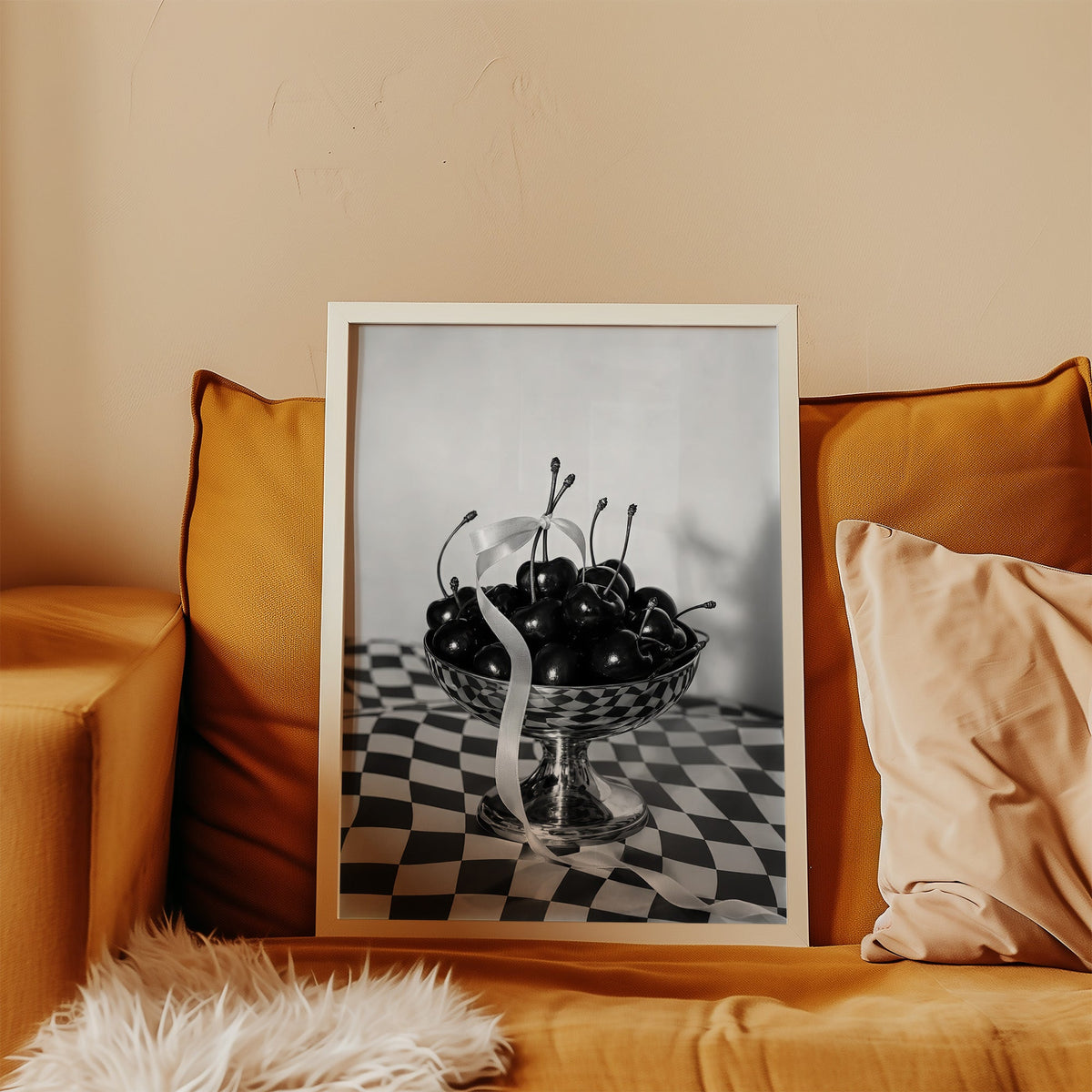 Black cherries in footed silver bowl print in a white frame leaning on a mustard velvet sofa