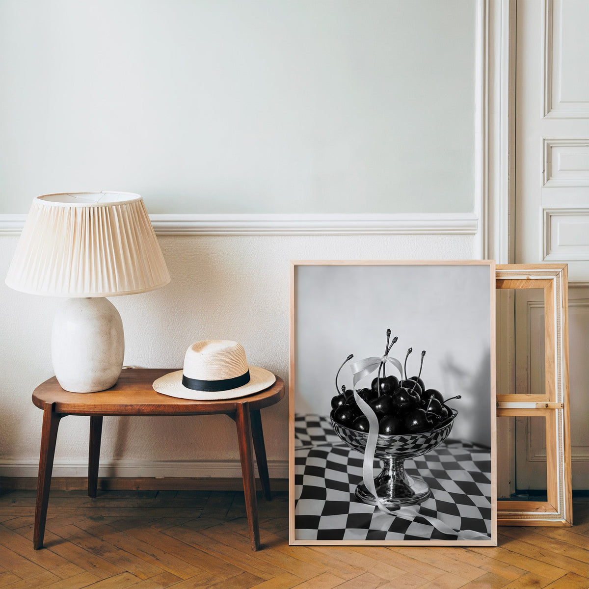 Black cherries in footed silver bowl print in a pine frame on a wood floor beside a lamp and straw hat