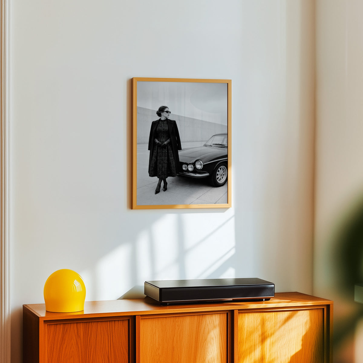Black and white woman in tweed coat by classic car print in an oak frame above a teak sideboard with a yellow glass lamp