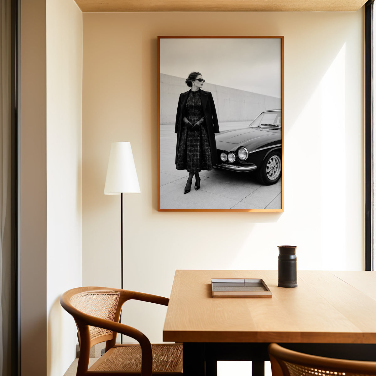 Black and white woman in tweed coat by classic car print in an oak frame above a dining table and cane chair