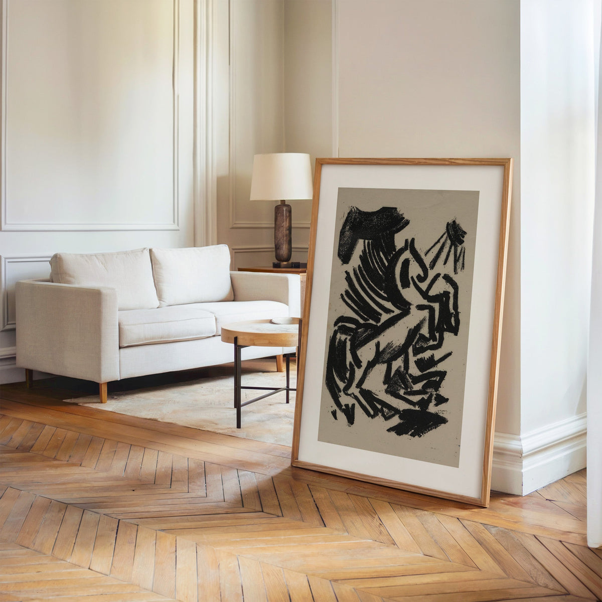 Black crayon rearing animal and sun print in an oak frame leaning on a herringbone floor by a cream linen sofa