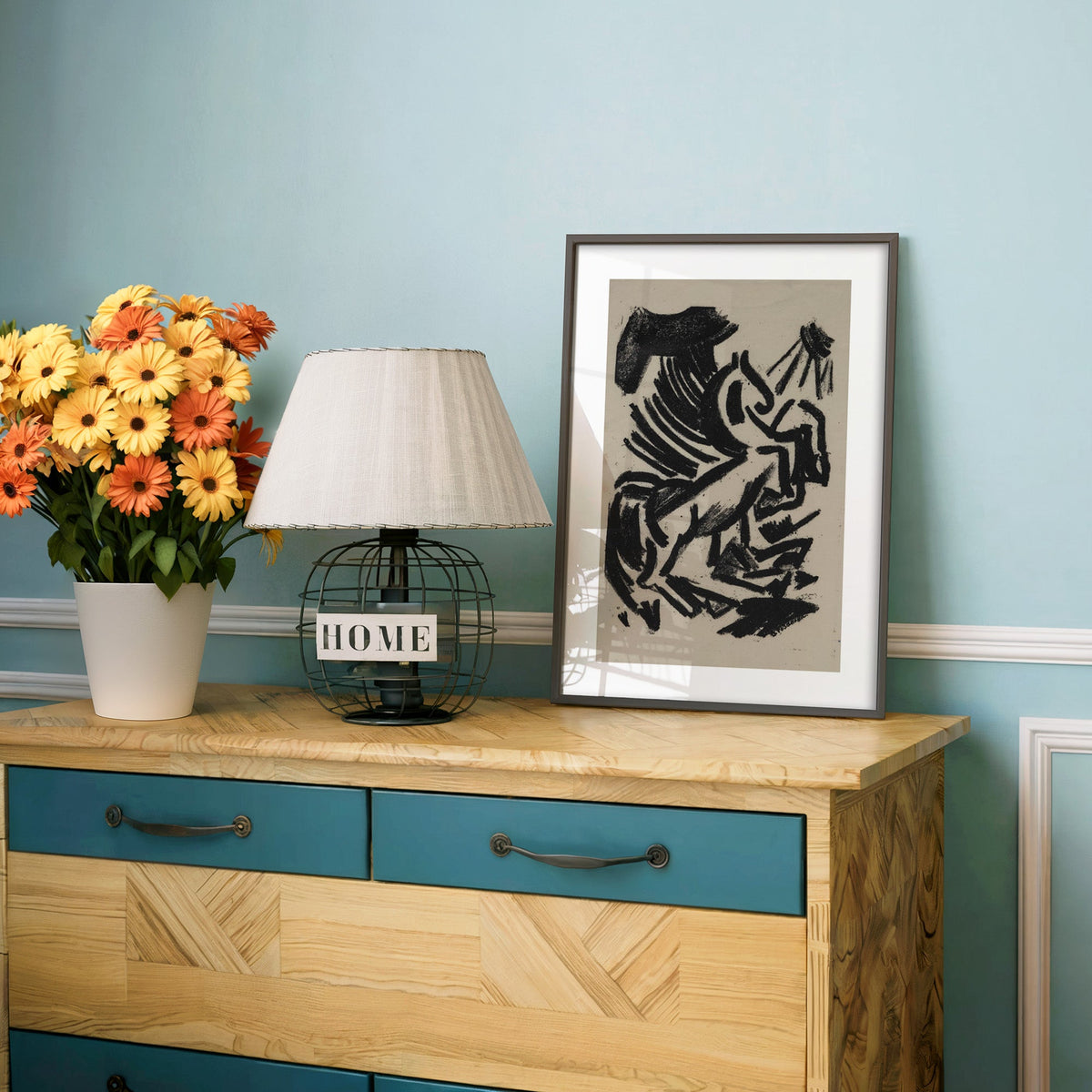 Black crayon rearing animal and sun print in a charcoal frame on a teal dresser beside orange and yellow daisies