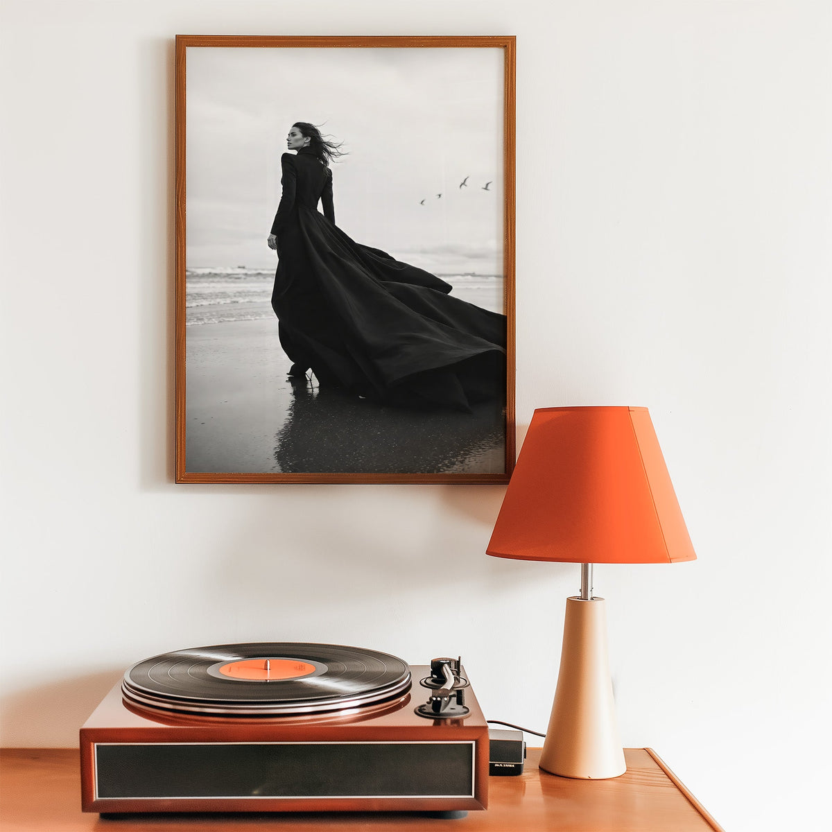 Woman in long black gown on a windy beach print in a walnut frame above a record player and orange lamp